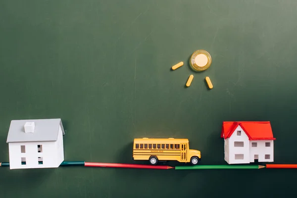 Top view of house models, toy school bus on road, made of color pencils, and sun made of magnets on green chalkboard — Stock Photo