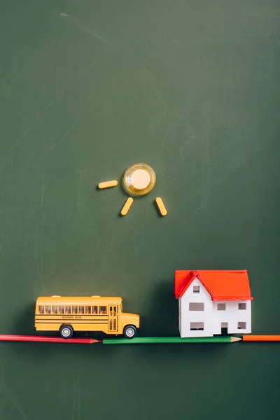 Top view of toy school bus on road made of color pencils near house model, and sun made of magnets on green chalkboard — Stock Photo