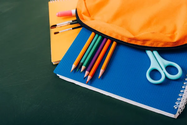 High angle view of backpack with notebooks, color pencils, paintbrushes and scissors on green chalkboard — Stock Photo