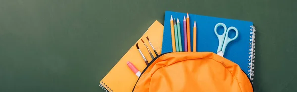 Imagem horizontal de cadernos, lápis de cor, tesoura e pincéis em mochila amarela sobre quadro-negro verde — Fotografia de Stock