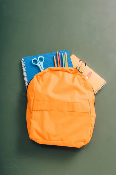 Top view of yellow backpack with copy books, scissors, color pencils and paintbrushes on green chalkboard — Stock Photo