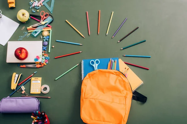 Top view of backpack with notebooks, color pencils and scissors near school supplies on green chalkboard — Stock Photo