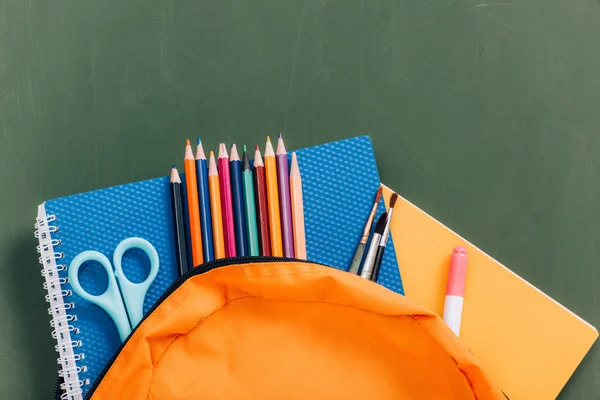 Top view of yellow backpack with color pencils, notebooks, scissors and paintbrushes near green chalkboard — Stock Photo