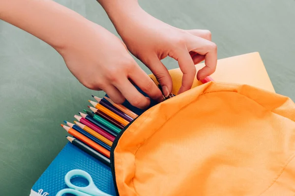 Vista recortada de los estudiantes tomando artículos de papelería de la mochila con cuadernos, lápices de color y tijeras en la superficie verde - foto de stock