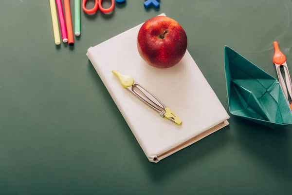 Vista dall'alto del gustoso divisore di mele e bussole sul libro vicino ai materiali scolastici sulla lavagna verde — Foto stock