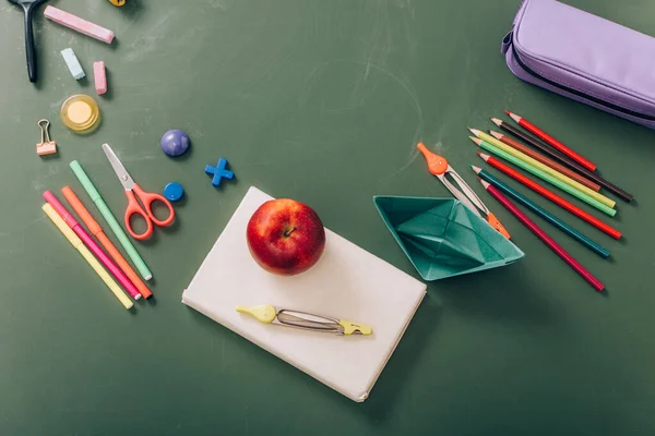 Vista dall'alto di mela matura e bussola divisorio su libro vicino barca di carta e materiale scolastico su lavagna verde — Foto stock