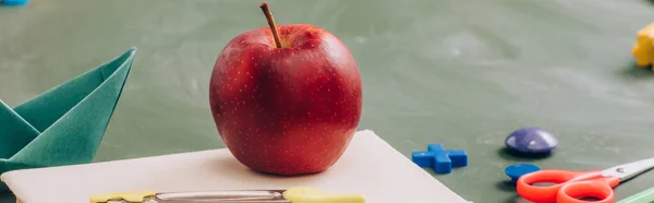 Enfoque selectivo de la manzana deliciosa en el libro cerca de papelería escolar en pizarra verde, concepto panorámico - foto de stock