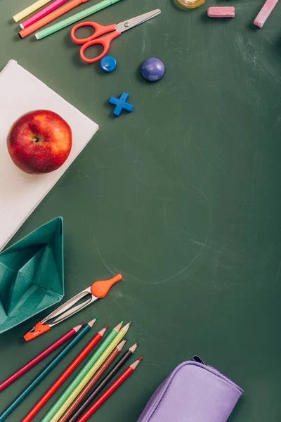 Top view of ripe apple on book, paper boat and school supplies on green chalkboard — Stock Photo
