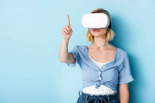 Young woman in virtual reality headset pointing with finger on blue — Stock Photo