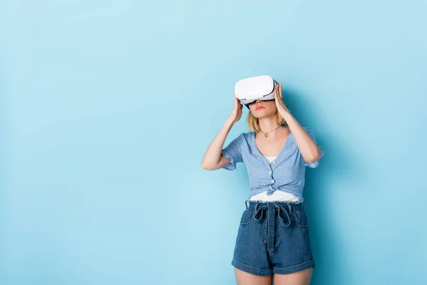 Junge Frau berührt Virtual-Reality-Headset und steht auf blau — Stockfoto