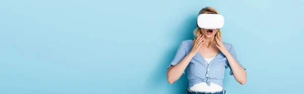 Panoramic crop of young and shocked woman in virtual reality headset on blue — Stock Photo