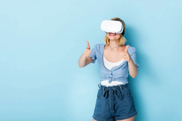 Молода жінка в гарнітурі віртуальної реальності показує великі пальці вгору на синьому — стокове фото