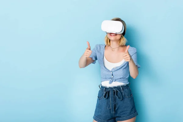 Giovane donna in realtà virtuale auricolare mostrando pollici in su sul blu — Stock Photo