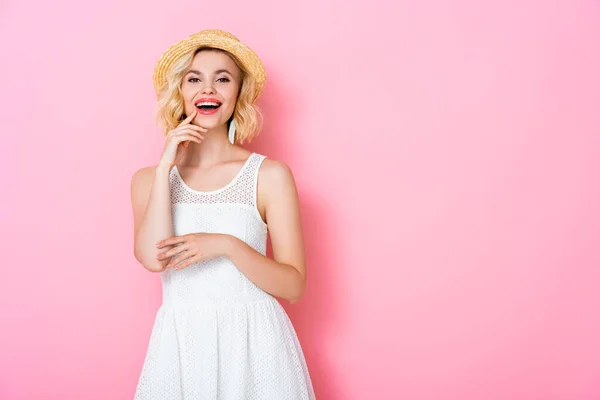 Excited young woman in white dress and straw hat touching face on pink — Stock Photo