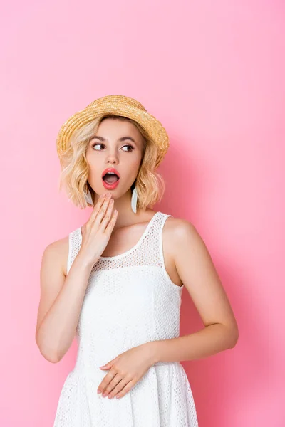Shocked young woman in white dress and straw hat looking away on pink — Stock Photo