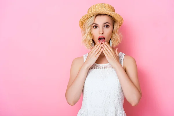 Shocked young woman in white dress and straw hat looking at camera and covering mouth on pink — Stock Photo