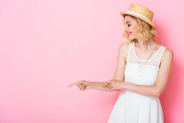 Mulher de vestido branco e chapéu de palha apontando com os dedos e olhando para longe em rosa — Fotografia de Stock