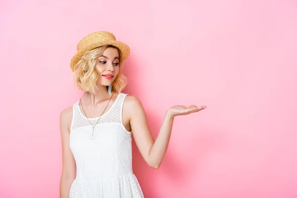 Mujer joven en sombrero de paja y vestido señalando con la mano en rosa - foto de stock