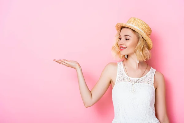 Mujer joven en sombrero de paja y vestido señalando con la mano en rosa - foto de stock