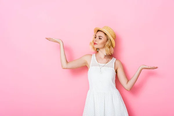 Mujer joven en sombrero de paja y vestido señalando con las manos en rosa - foto de stock