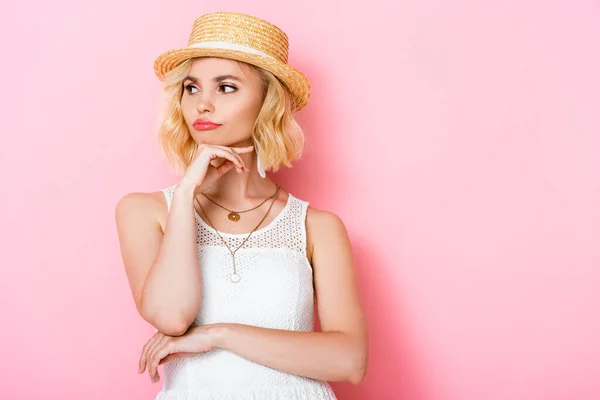 Pensive woman in straw hat touching face and looking away on pink — Stock Photo