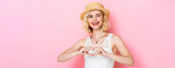 Panoramaaufnahme einer Frau mit Strohhut, zwinkerndem Auge und Herzzeichen mit Händen auf rosa — Stockfoto