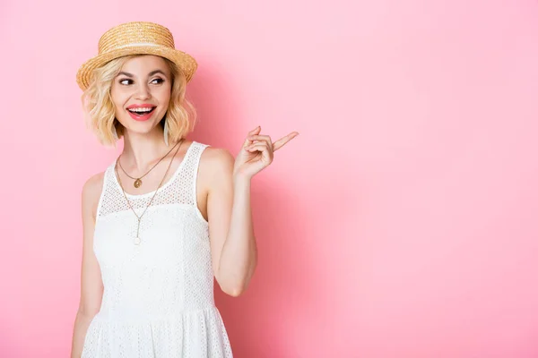 Mujer excitada en sombrero de paja señalando con el dedo mientras mira hacia otro lado en rosa - foto de stock