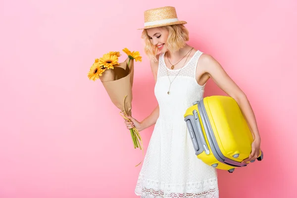 Young woman in straw hat holding yellow flowers and luggage on pink — Stock Photo