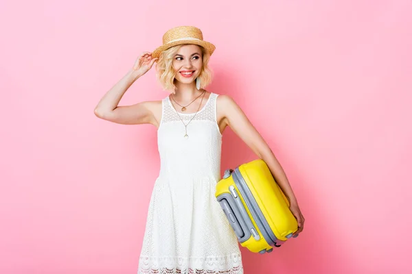 Young woman touching straw hat while holding yellow luggage on pink — Stock Photo