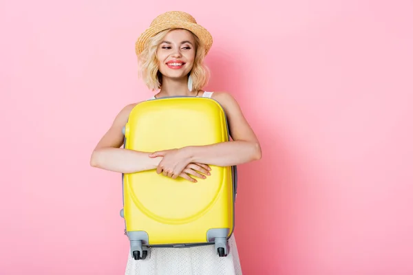 Woman in straw hat hugging yellow luggage on pink — Stock Photo