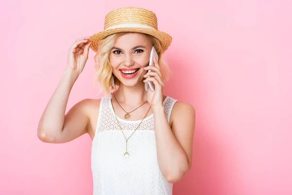 Mujer tocando sombrero de paja y hablando en el teléfono inteligente en rosa - foto de stock