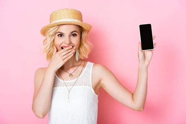 Donna in cappello di paglia che tiene smartphone con schermo bianco e bocca di copertura su rosa — Foto stock