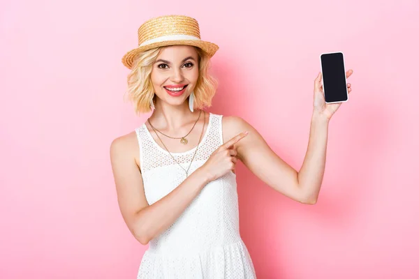 Mujer en sombrero de paja señalando con el dedo en el teléfono inteligente con pantalla en blanco en rosa - foto de stock
