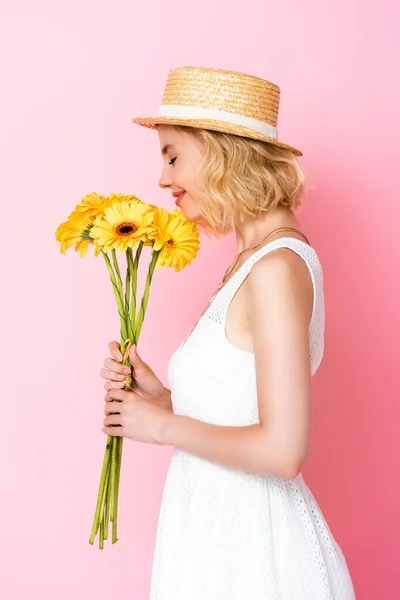 Вид сбоку женщины в соломенной шляпе и белом платье, пахнущем желтыми цветами на розовом — стоковое фото
