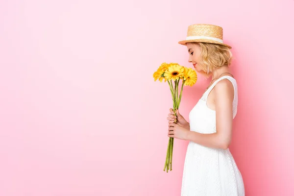 Vista lateral da mulher em chapéu de palha e vestido branco cheirando flores amarelas em rosa — Fotografia de Stock