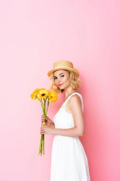 Young woman in straw hat and dress holding yellow flowers on pink — Stock Photo