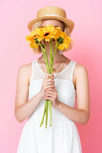 Young woman in straw hat and dress covering face with yellow flowers on pink — Stock Photo