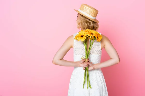 Vista posterior de la mujer joven en sombrero de paja y vestido blanco sosteniendo flores amarillas detrás de la espalda en rosa - foto de stock