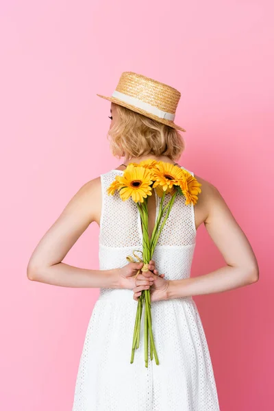 Back view of woman in straw hat and white dress holding yellow flowers behind back on pink — Stock Photo