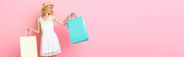 Panoramic shot of woman in straw hat and white dress holding shopping bags on pink — Stock Photo