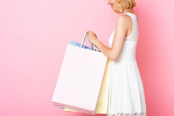 Cropped view of young woman in white dress holding shopping bags on pink — Stock Photo