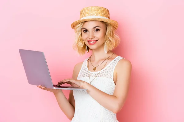 Жінка в солом'яному капелюсі використовує ноутбук на рожевому — стокове фото