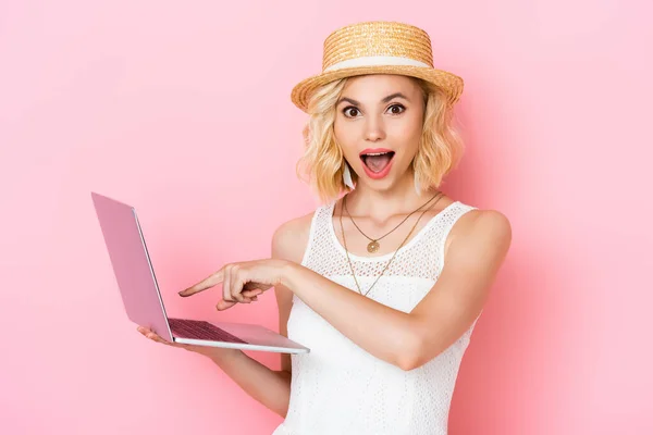 Femme excitée dans chapeau de paille pointant avec le doigt à l'ordinateur portable sur rose — Photo de stock