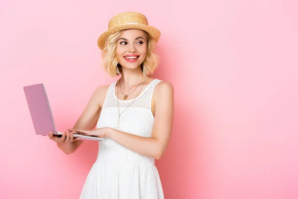 Giovane donna in cappello di paglia che tiene il computer portatile e distoglie lo sguardo sul rosa — Foto stock