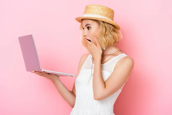 Mujer conmocionada en sombrero de paja mirando a la computadora portátil y la boca cubierta de rosa - foto de stock