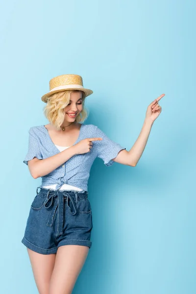 Woman in straw hat pointing with fingers and looking away on blue — Stock Photo