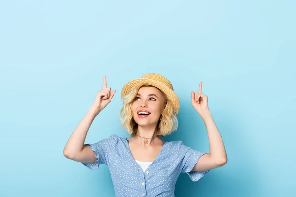 Young woman in straw hat pointing with fingers and looking up on blue — Stock Photo