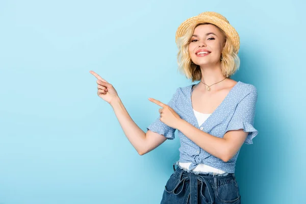 Junge Frau mit Strohhut zeigt mit den Fingern und blickt in die Kamera auf blau — Stockfoto