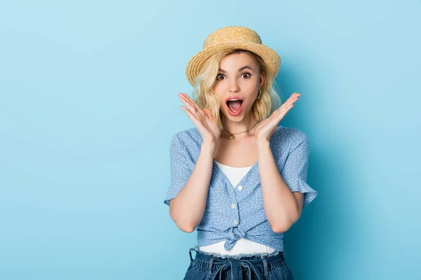 Shocked young woman in straw hat gesturing and looking at camera on blue — Stock Photo