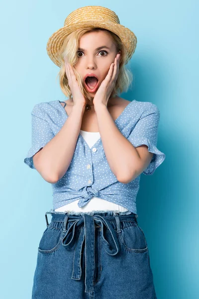 Shocked young woman in straw hat touching face and looking at camera on blue — Stock Photo